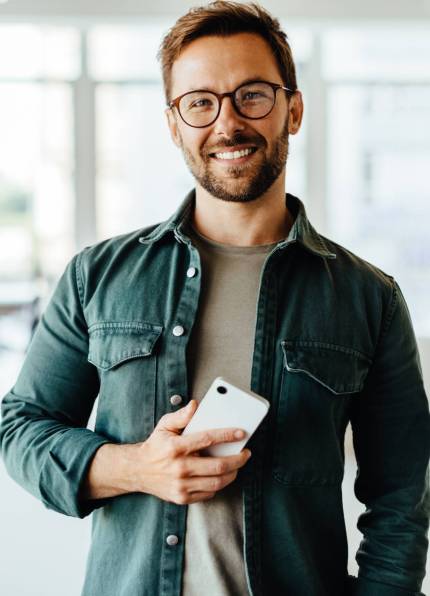 Portrait of a male professional standing in an office Portrait of a male professional standing in an office with his colleagues in the background. Business man looking at the camera while holding a mobile phone.